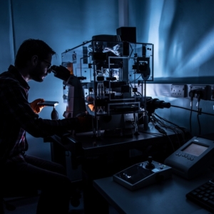 A man silhouetted in dim blue light peers into a microscope beside a complex piece of lab equipment illuminated by internal lighting.