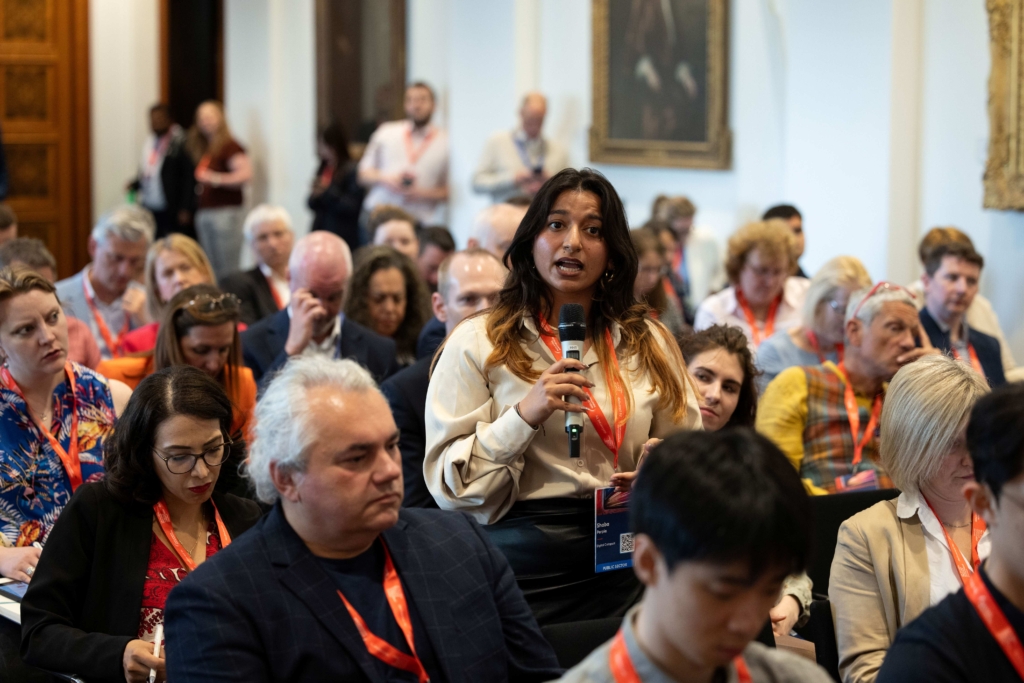 Image of a person stood up talking, with a microphone in an audience of seated people
